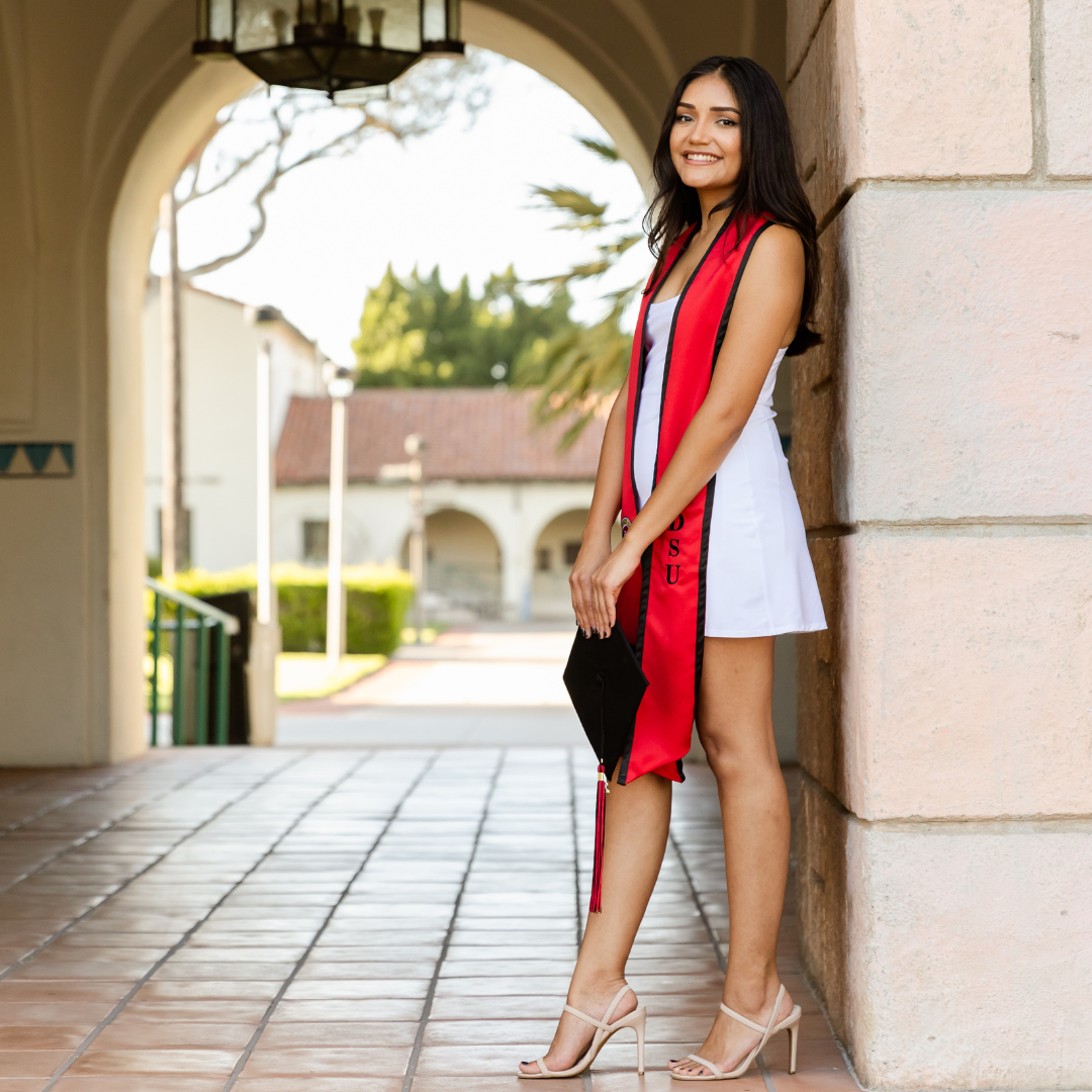 Graduate wearing white dress and red graduation stole poses elegantly under an arched walkway on campus.