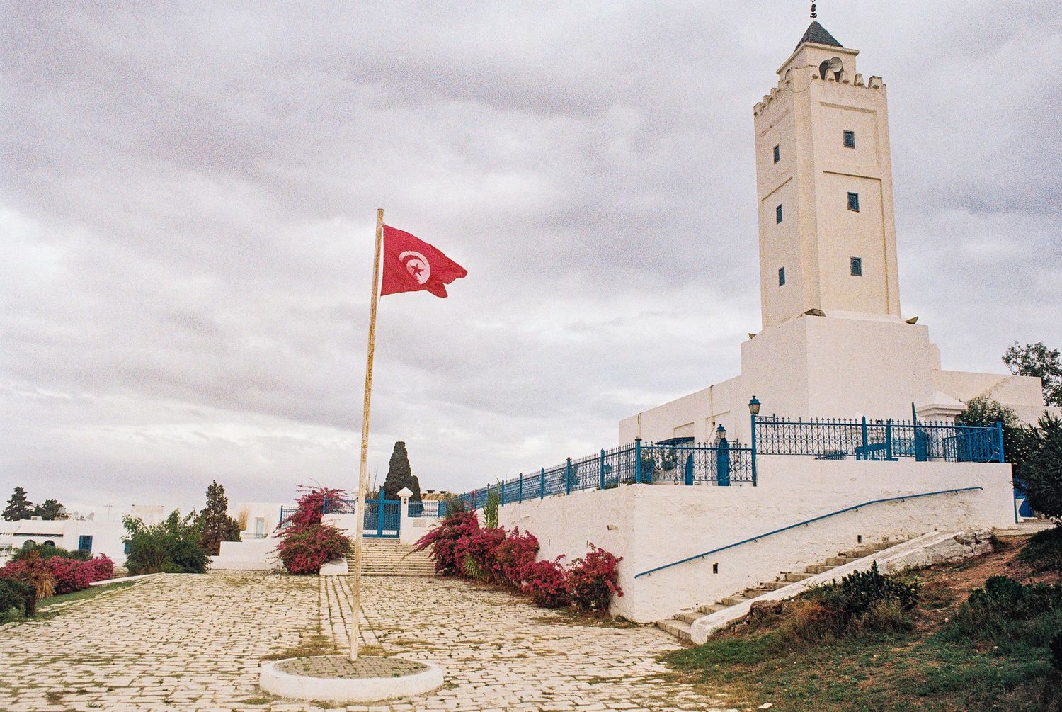 White lighthouse tower with Tunisian flag flying on hilltop under cloudy skies in coastal Mediterranean setting.