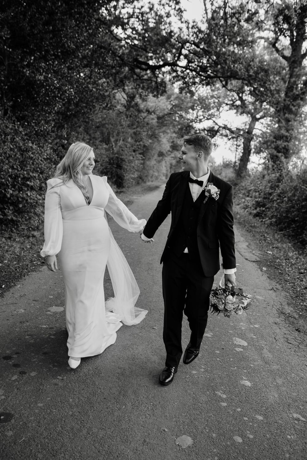 A newly married couple take a romantic joyful walk down a countryside lane, holding hands. The groom holds the bouquet.