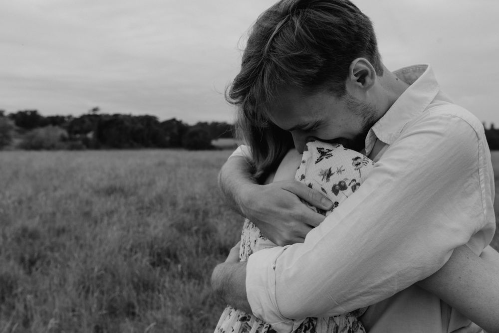 A loving couple hug deeply in a field out in the countryside. The woman wears a ditsy floral dress.