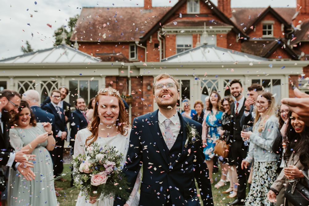 A bride and groom walk through a shower of confetti after their ceremony. The groom is wearing a blue suit.