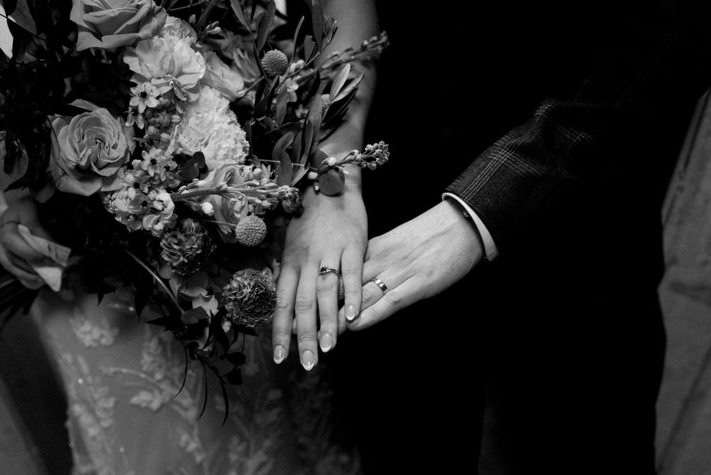 Black and white close-up of hands displaying wedding rings next to an elegant floral bouquet.