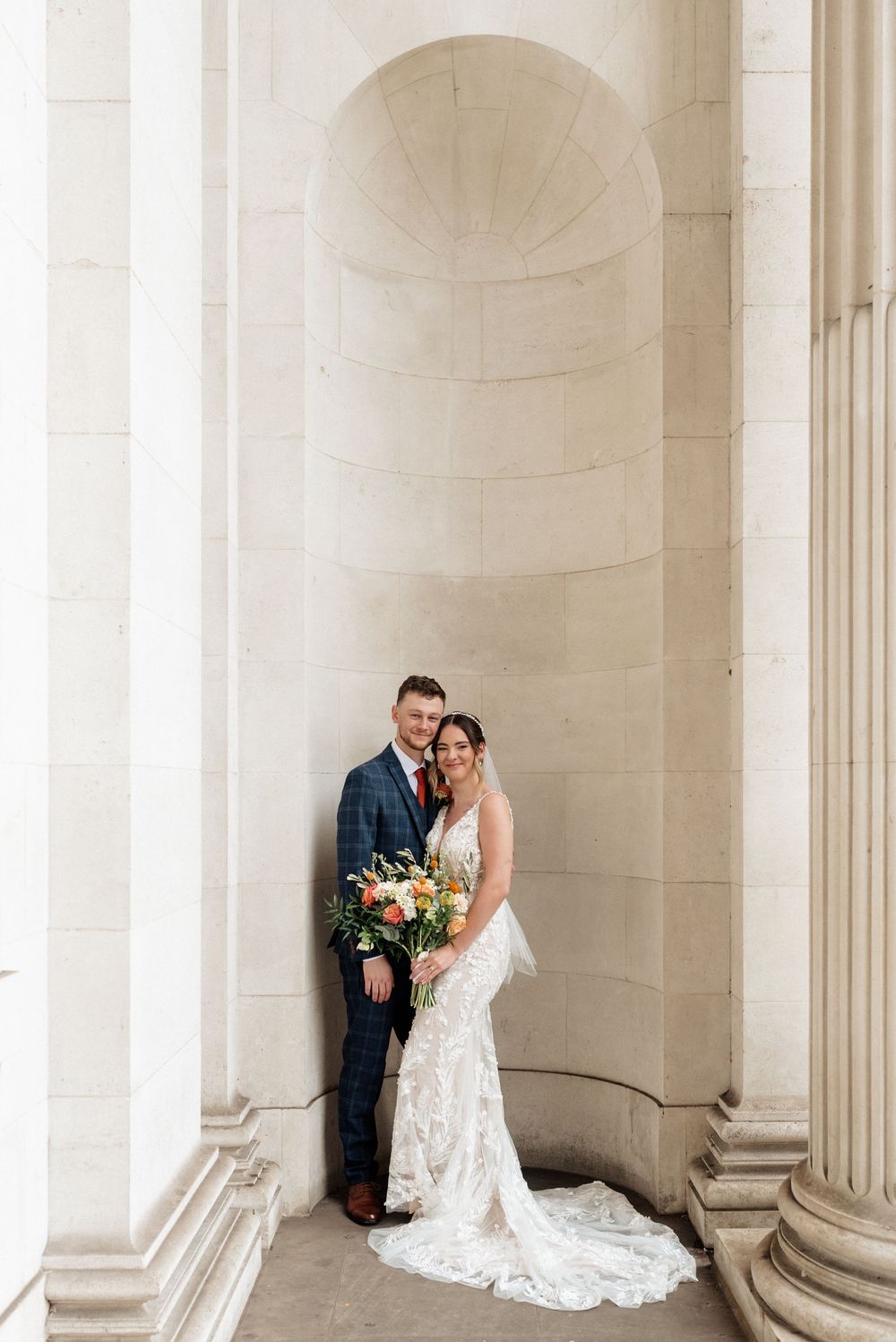 Elegant wedding couple poses between classic white marble columns in architectural setting with flowing bridal gown.