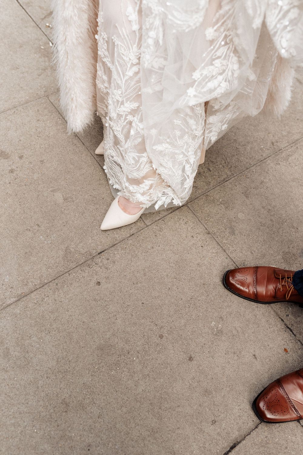 Close-up of wedding shoes showing white bridal heels and brown leather groom's shoes on concrete.