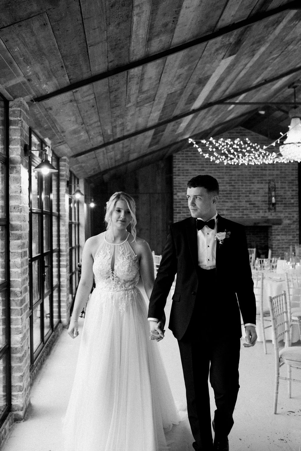 A black and white photo of a bride and groom walking hand in hand down a covered brick pathway decorated with lights.