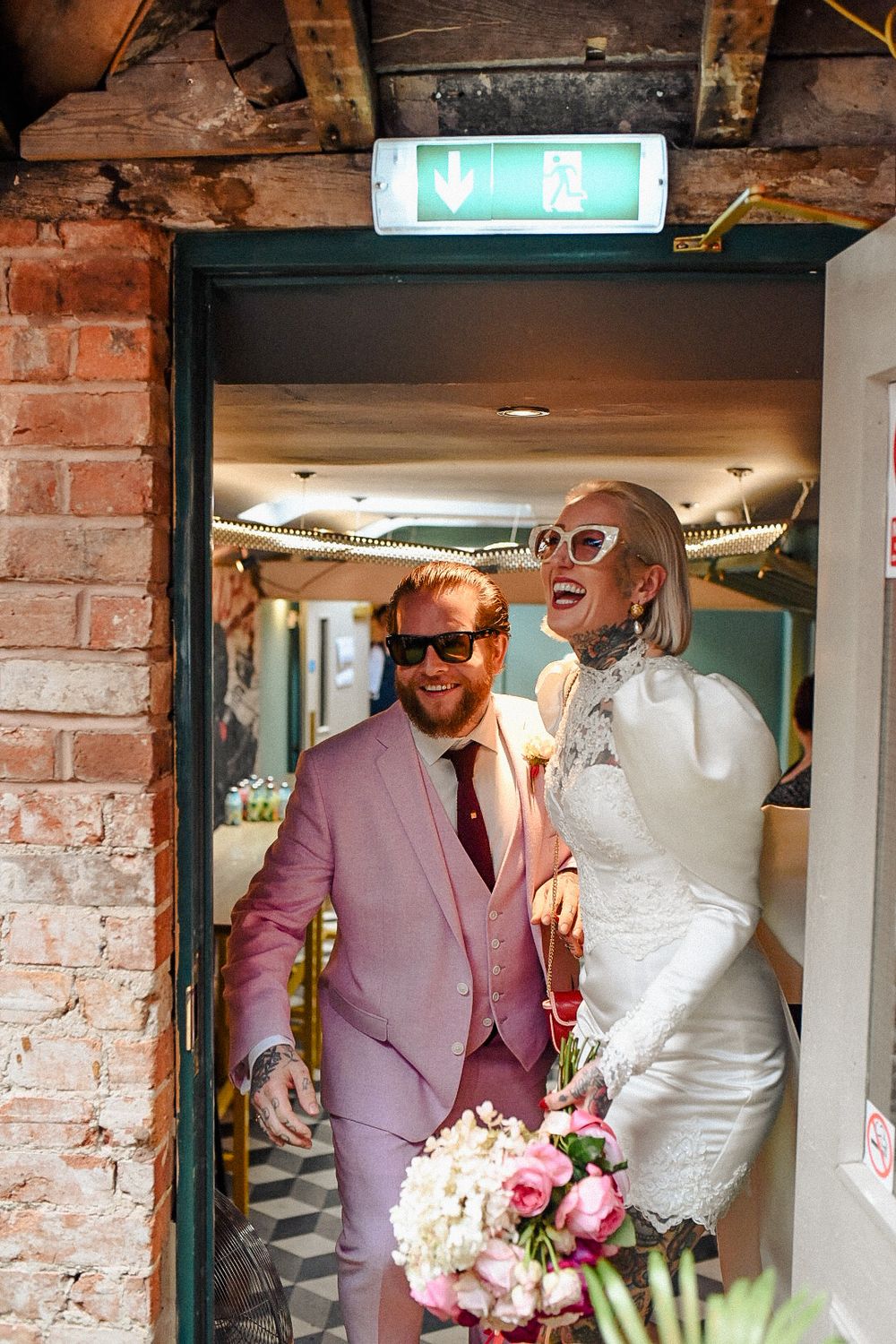 A newly wed tattooed couple enter their wedding reception wearing sunglasses. Groom wears a pink suit.
