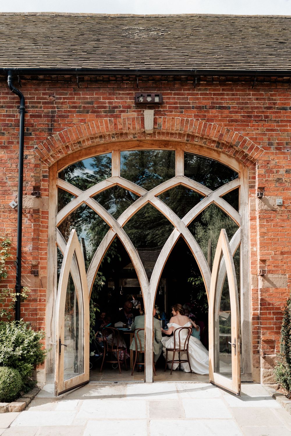 A bride and groom sit down at their wedding breakfast inside the open doors of a large barn conversion wedding venue.