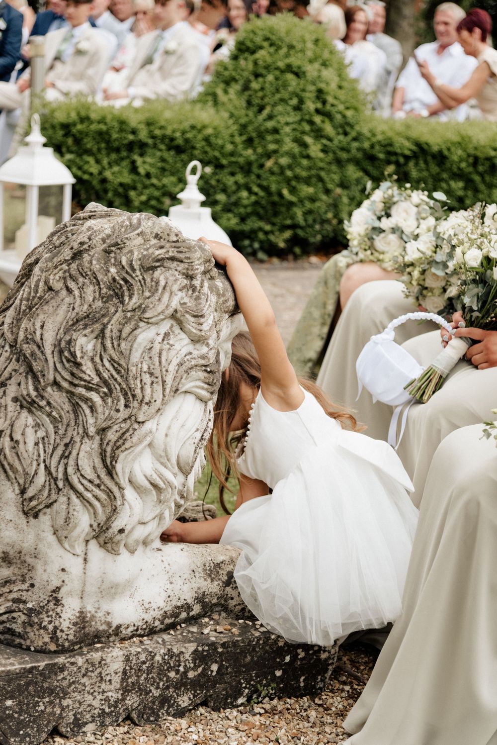 A young flower girl explores a stone lion statue at an outdoor wedding ceremony.