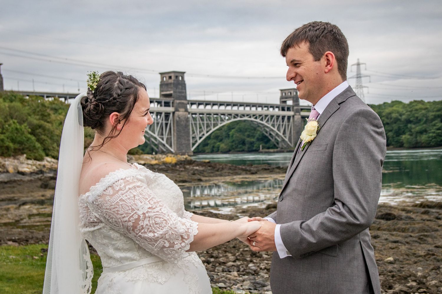 A wedding portrait of a couple looking at each other holding hands with Britannia bridge behind. Anglesey Wedding Photographer.