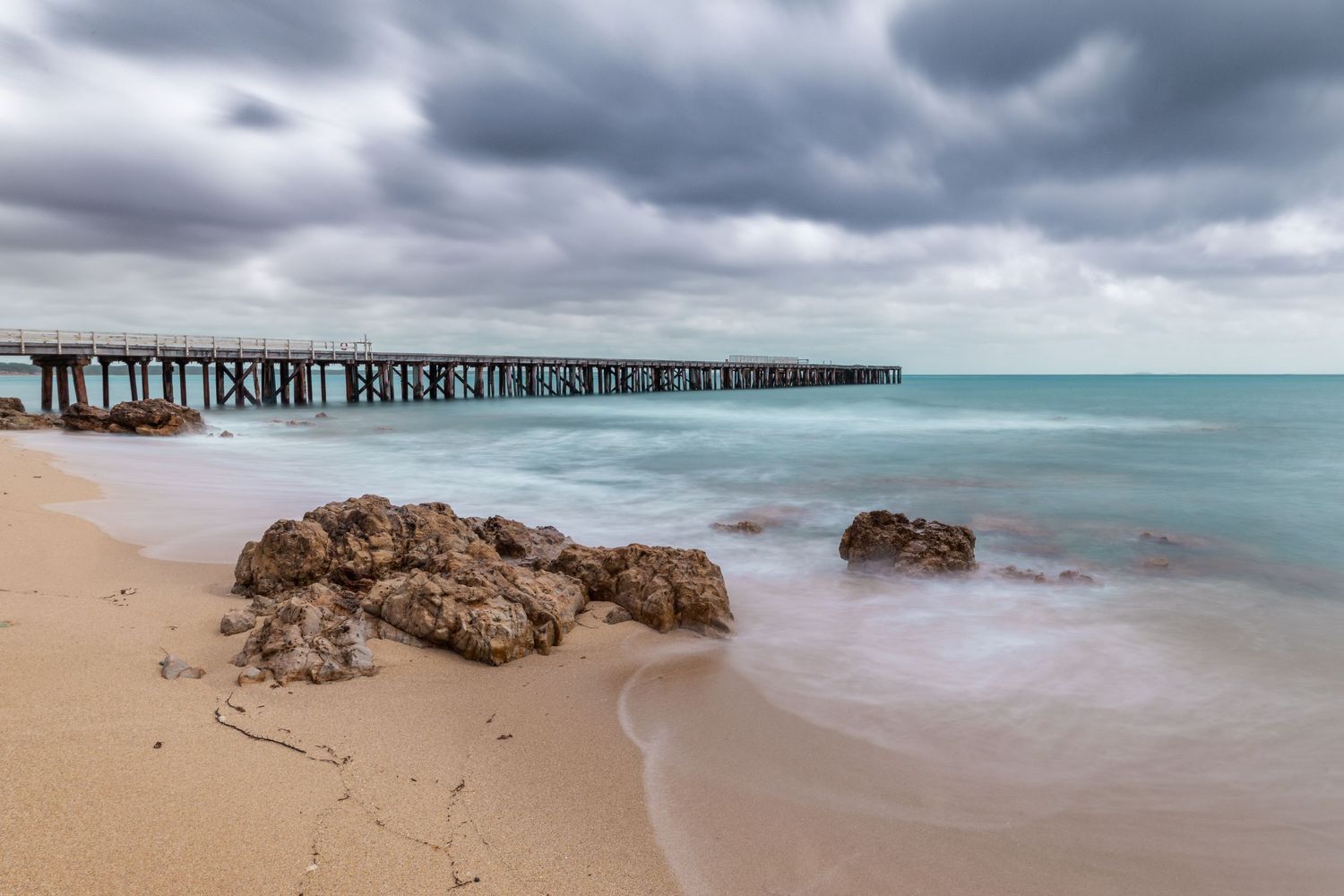 Long wooden pier extends into turquoise ocean under moody storm clouds.