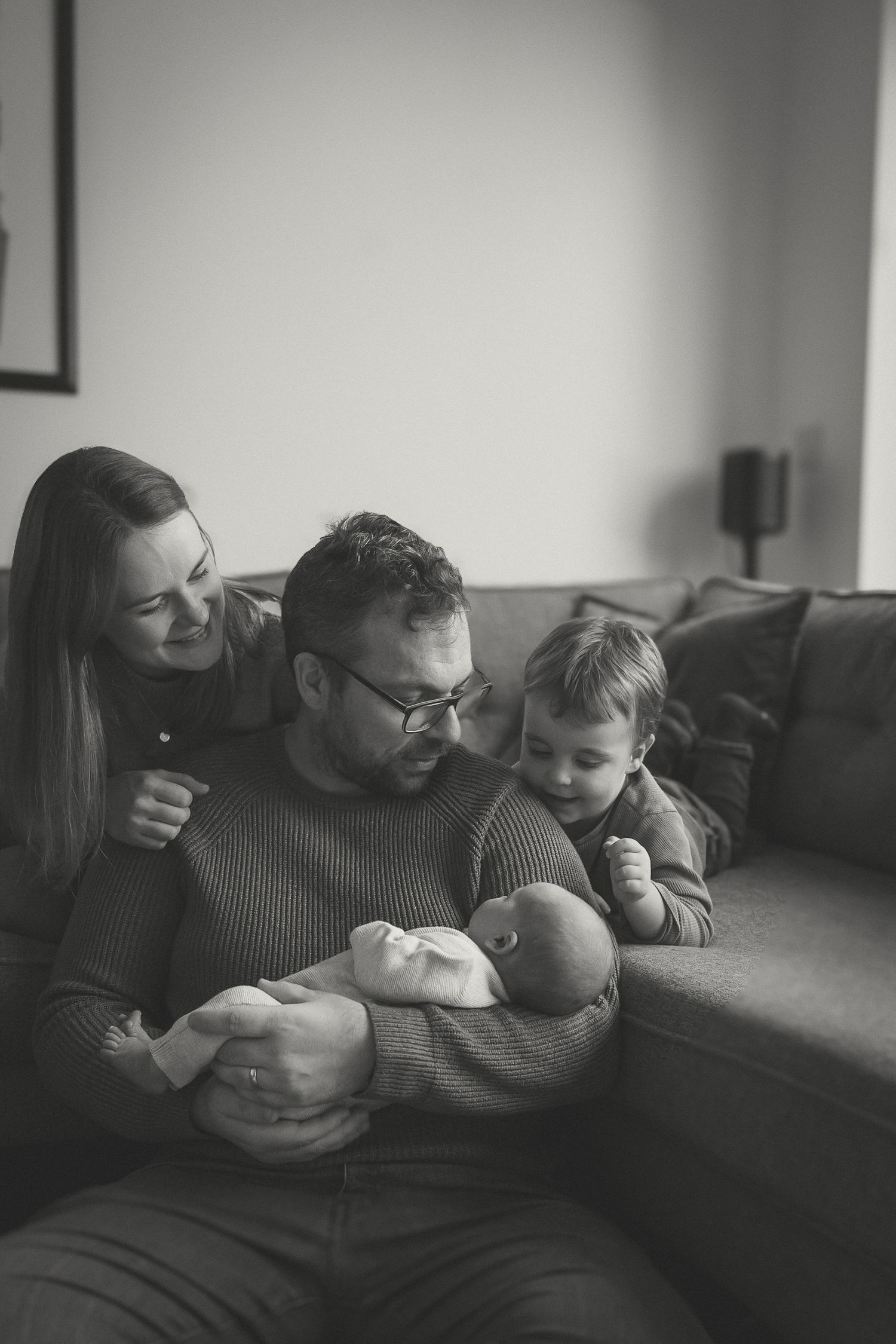 A black & white image of a family cuddling together on a sofa while holding their newborn.