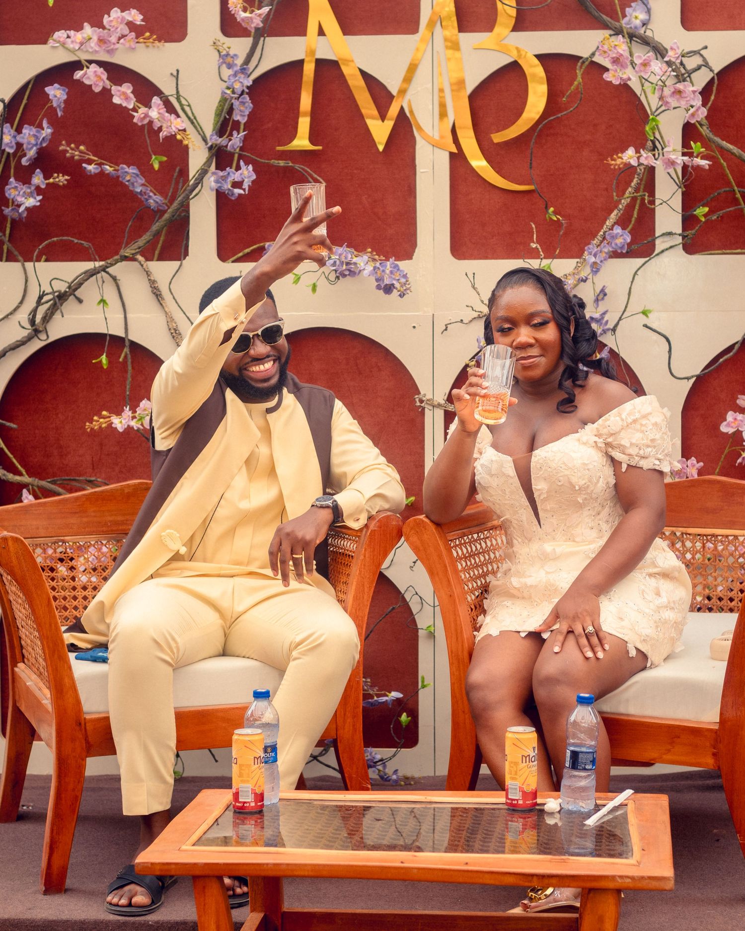 Couple in cream and white attire share drinks while sitting on wooden chairs.