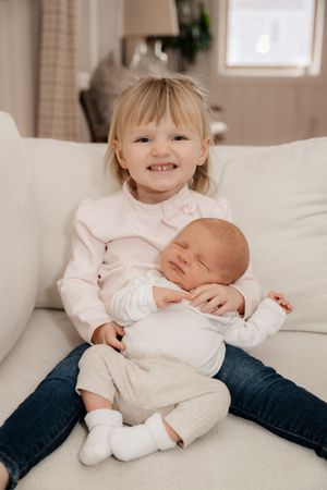 Two children sit together on a white couch, one cradling a baby in a light-colored outfit. Southern Maine Newborn Lifestyle