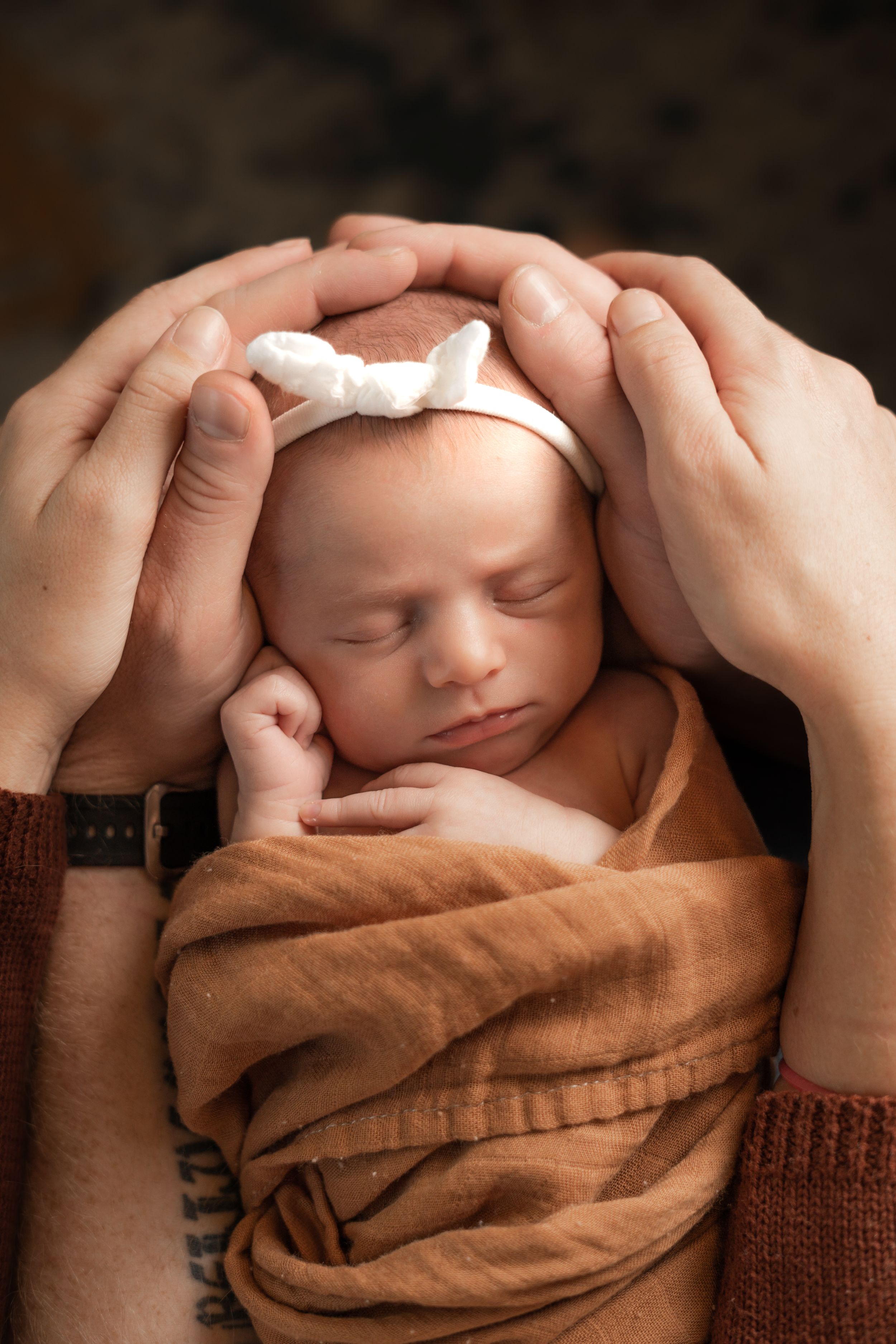Sleeping newborn wrapped in brown blanket with hands framing face.