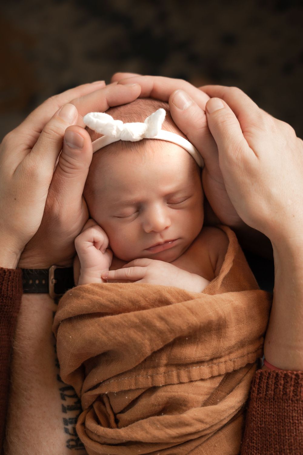 Sleeping newborn wrapped in brown blanket with hands framing face.