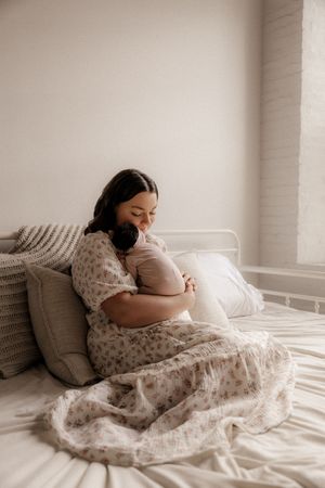 Pregnant woman in floral dress sitting on white bed, cradling her baby bump with a serene smile.