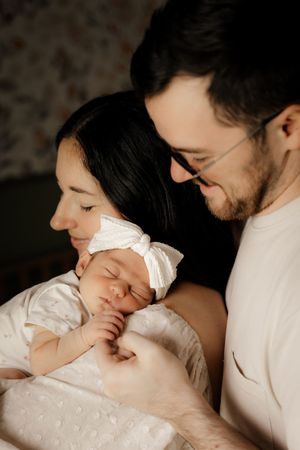New parents lovingly gazing at their sleeping newborn baby girl wearing a white bow headband and outfit.