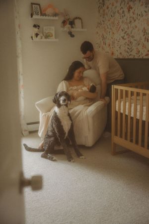 A couple admires their newborn in a nursery glider while their curly-haired dog sits attentively nearby.