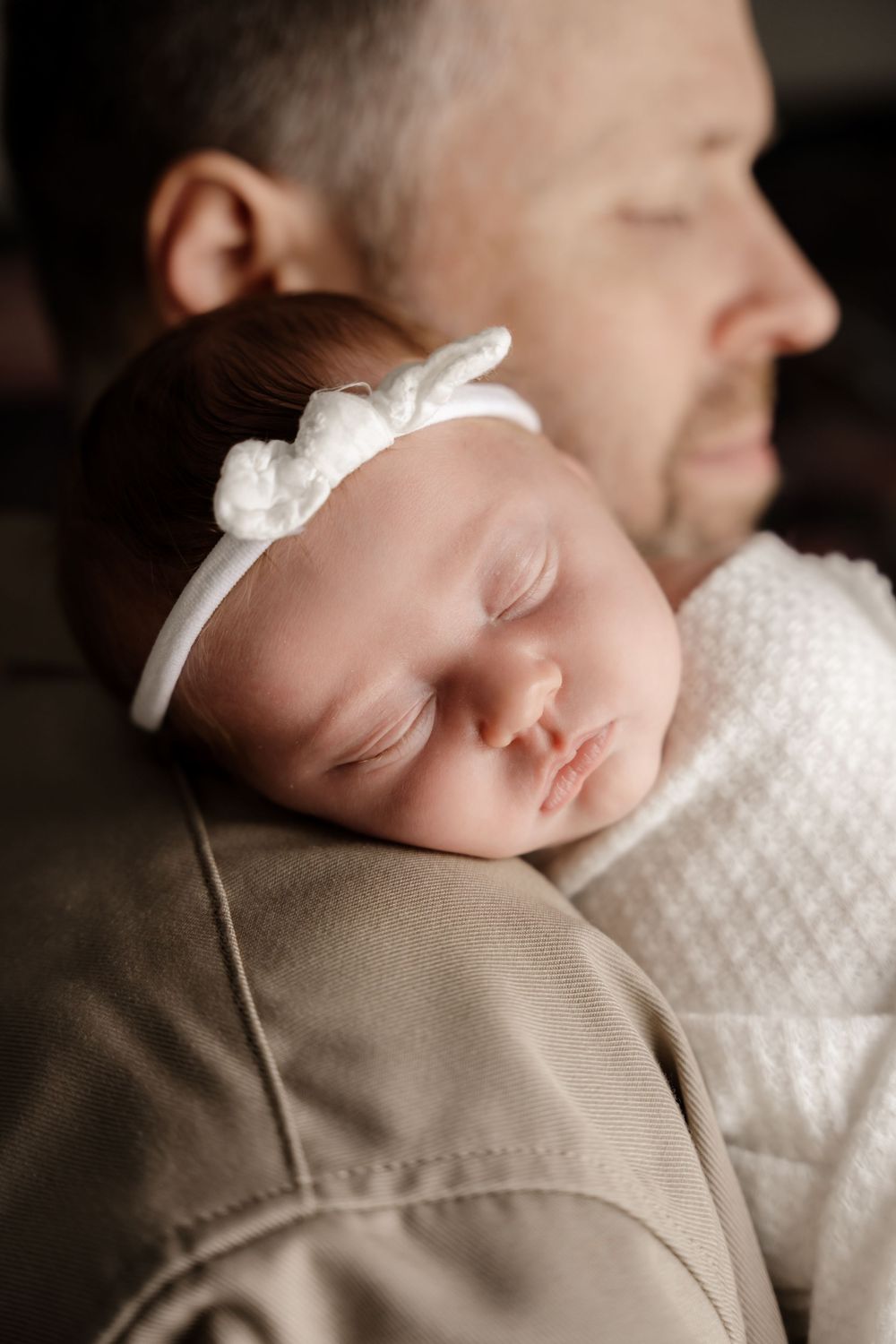 Sleeping newborn wearing white with a small bow accessory resting on beige fabric.