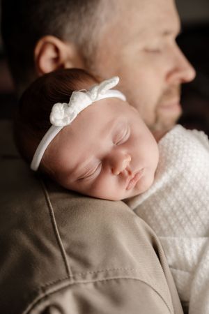 Sleeping newborn wearing white with a small bow accessory resting on beige fabric.