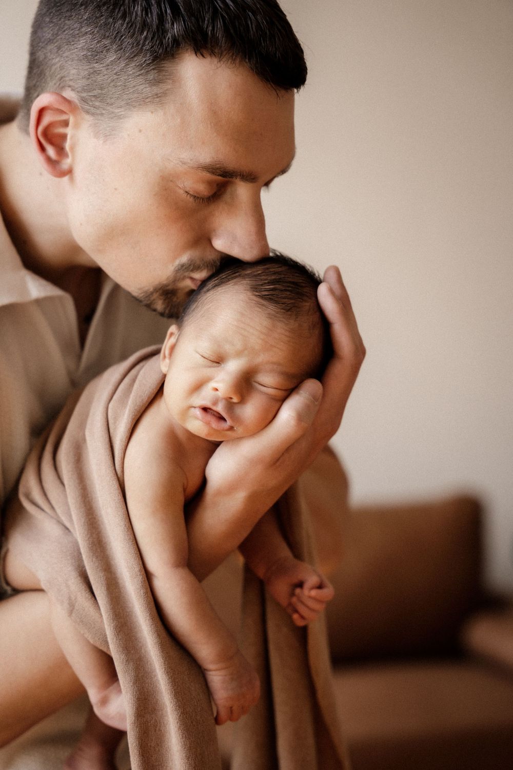 A father tenderly kisses his sleeping newborn baby wrapped in a soft beige blanket.
