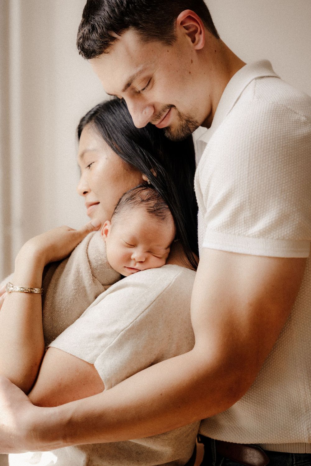 A tender family portrait of parents cradling their sleeping newborn baby, captured in soft neutral tones.