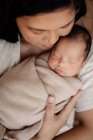A mother tenderly kisses her sleeping newborn baby wrapped in a soft beige swaddle blanket.