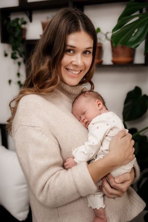 Parent in cream sweater holding and bonding with newborn baby against white wall with plants.