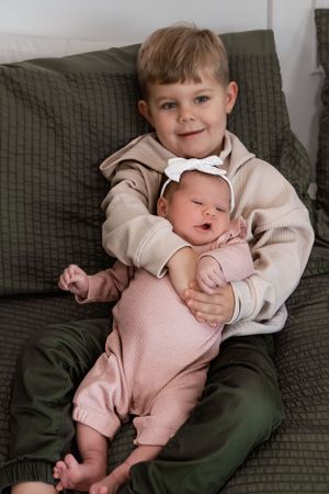 Young child in beige sweater holding baby sibling on green couch.