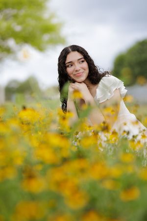 Young woman with curly dark hair in white dress smiling dreamily among vibrant yellow wildflowers in a lush green field.