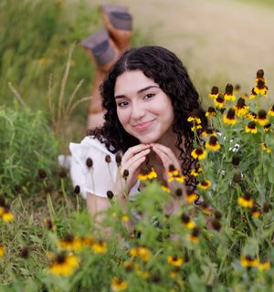Young woman with curly hair lying among yellow black-eyed Susan wildflowers in a lush green field, smiling warmly.