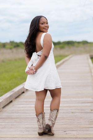 Young woman in white dress and cowboy boots posing on wooden boardwalk surrounded by green fields.