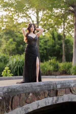 Woman in black beaded gown with slit poses on stone bridge surrounded by lush green trees.