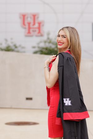 Smiling woman in red dress holding UH graduation gown outside University of Houston campus.