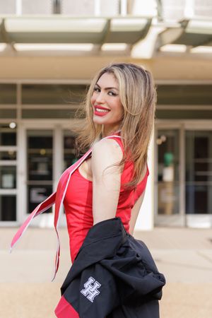 Smiling blonde woman in red top holding graduation gown outside University of Houston building.