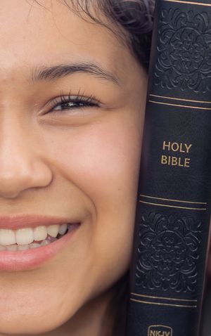 Close-up portrait of a warm smile against a dark background with a Bible book spine visible.
