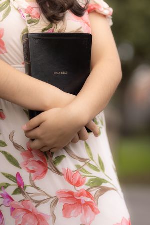 Someone in a floral dress holds a black Bible or holy book close to their chest in an outdoor setting.