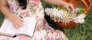 Person in floral dress sits in grass with wicker basket and daisies nearby while writing in journal. In Pearland, TX