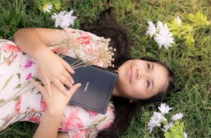 Person in a floral dress lying in grass surrounded by scattered white flowers on a sunny day. In Pearland, TX