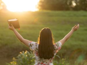 A person in a floral dress stands with raised arms holding a bible against a golden sunset in a grassy field. In Pearland, TX