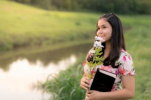 A person in black top holds a blooming pink flower while standing in a sunlit outdoor natural setting. In Pearland, TX