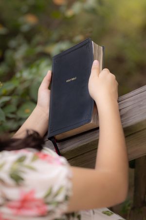 Hands holding a black Holy Bible while sitting on a wooden deck outdoors with blurred foliage in background.