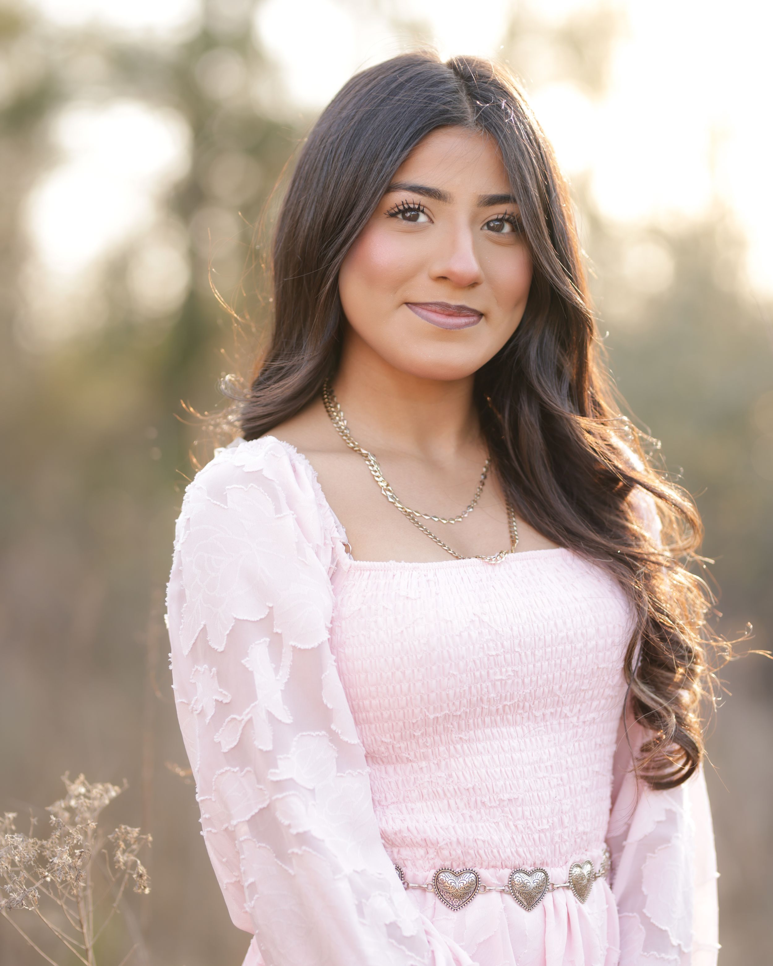 Young woman in pink smocked dress and ruffled cardigan with heart belt buckle poses outdoors in golden hour light.