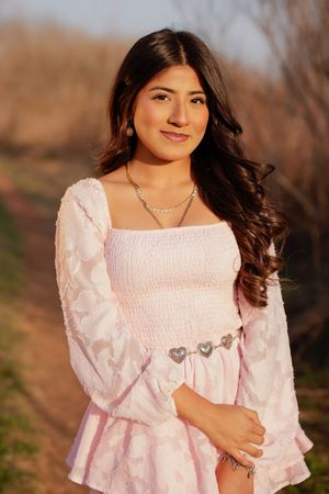 Young woman in pink smocked dress with heart belt, silver necklace, posing outdoors in warm sunset light.