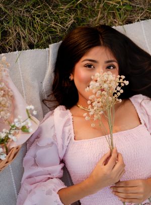 Young woman in pink smocked dress lying on blanket outdoors, holding delicate white baby's breath flowers near her face.