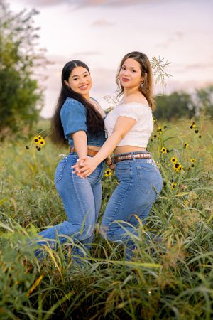 Two friends in blue jeans pose playfully among wildflowers during a golden hour photoshoot.