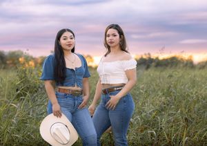 Two people in denim sit together in a grassy field at sunset holding a straw hat.