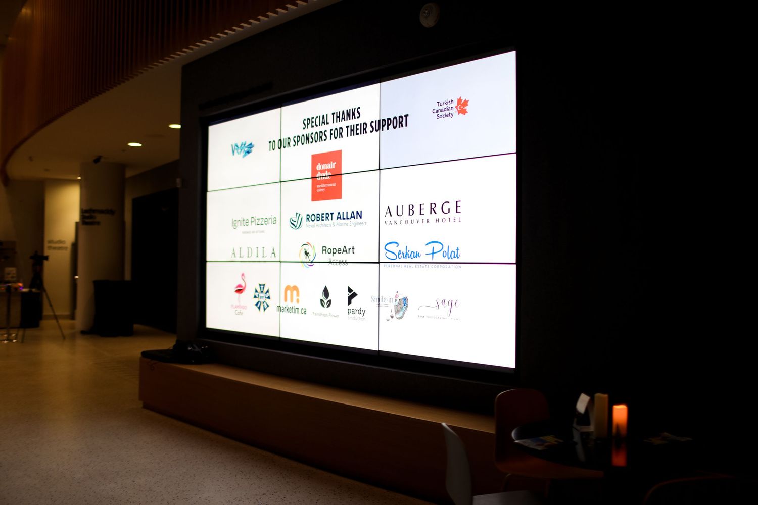 Large illuminated display board showing multiple corporate sponsor logos and event branding in a dark interior space.