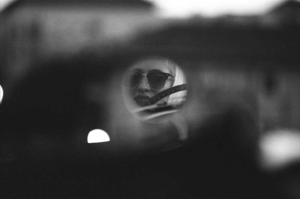 Black and white reflection of a person wearing sunglasses captured in a car mirror.