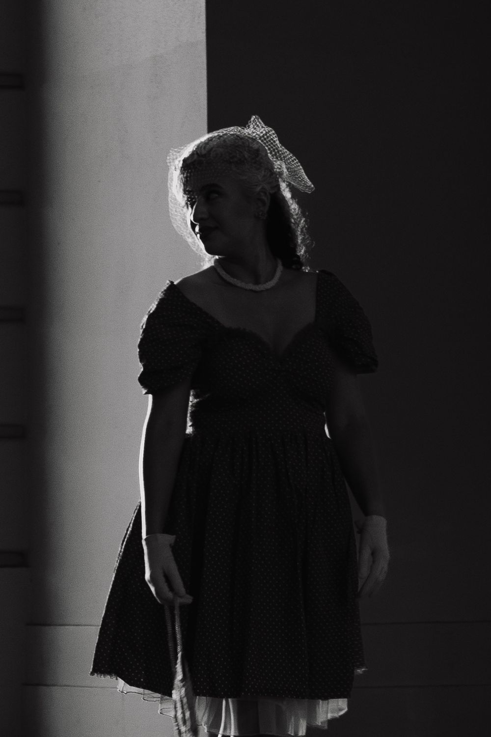 Black and white photo of a young girl in a polka dot dress and hair bow standing in dramatic light.